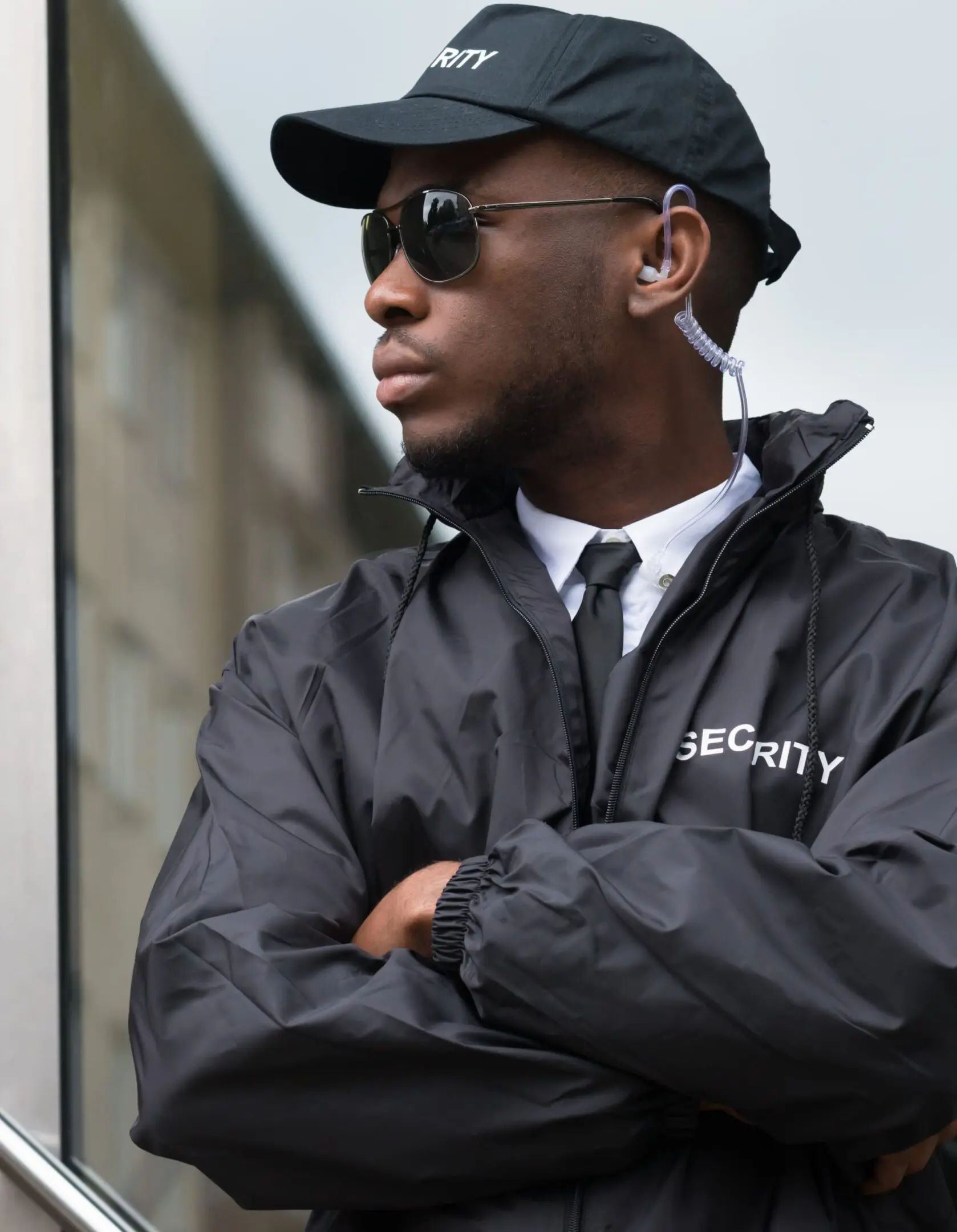 Frame 1618873616 (1) A security guard wearing sunglasses and a black uniform with "Security" written on the cap and jacket stands with crossed arms in front of a building.