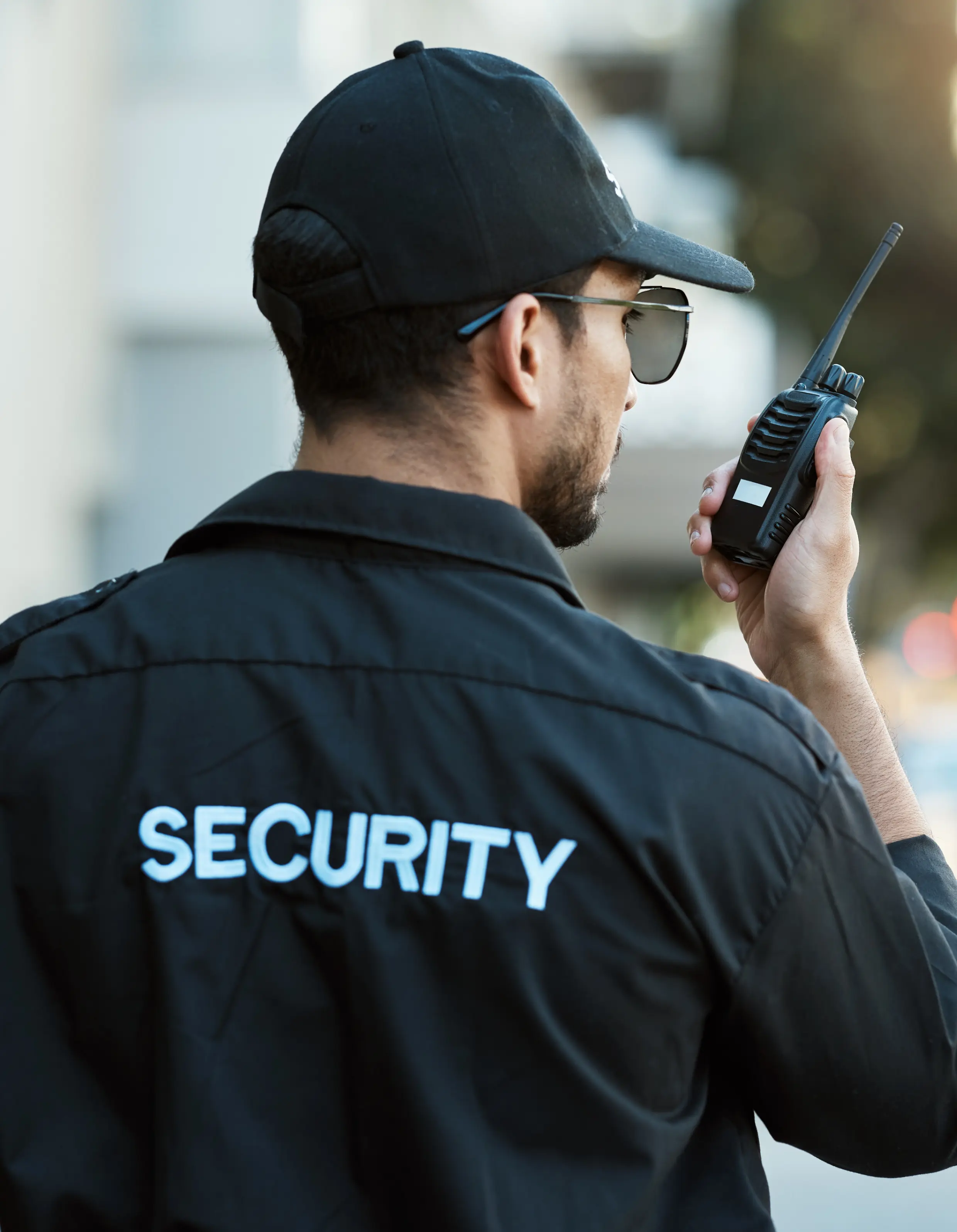 Frame 1618873616 A person wearing a black security uniform and cap is holding a walkie-talkie, looking away. The word "SECURITY" is visible on the back of the jacket.