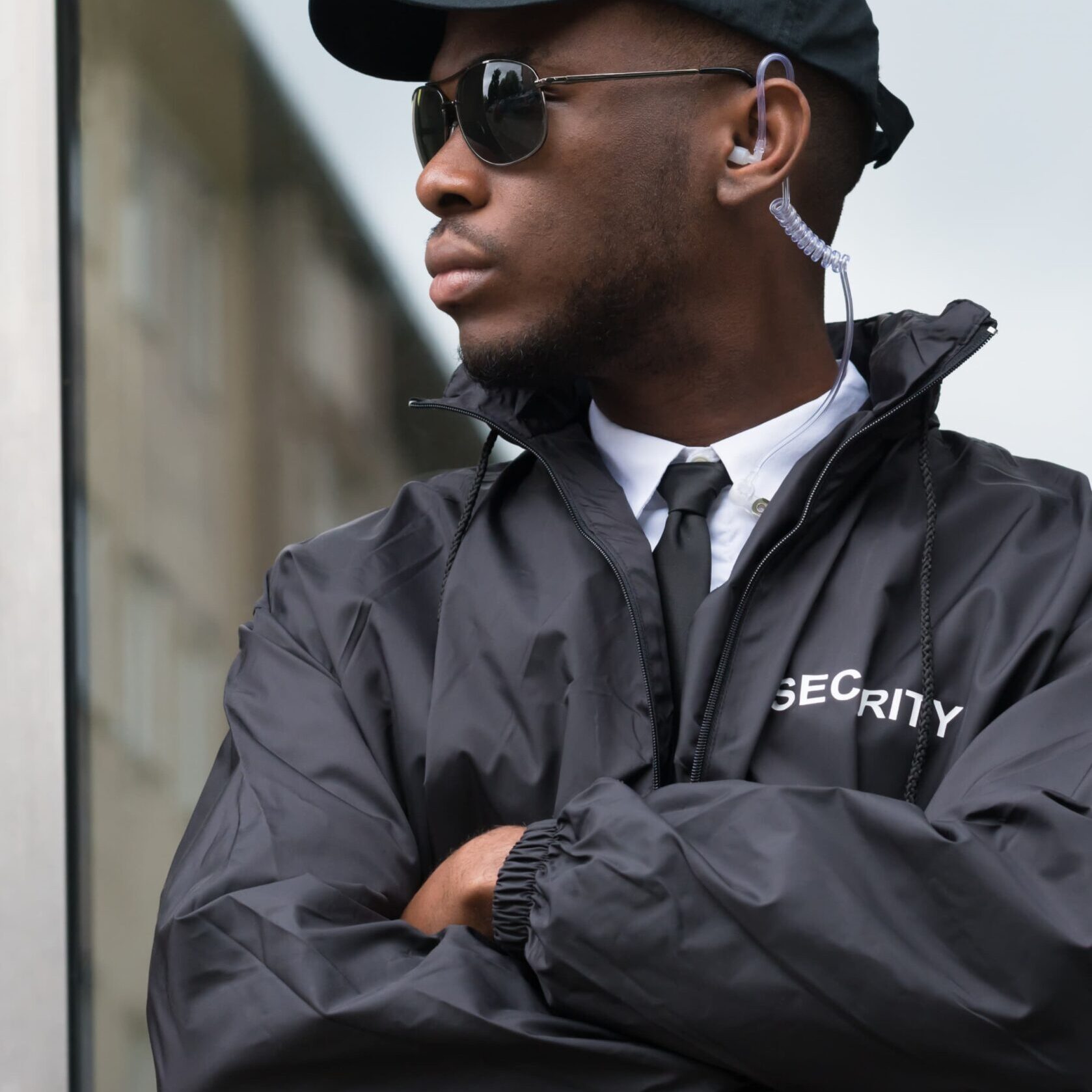 Frame 1618873616 (1) A security guard wearing sunglasses and a black uniform with "Security" written on the cap and jacket stands with crossed arms in front of a building.